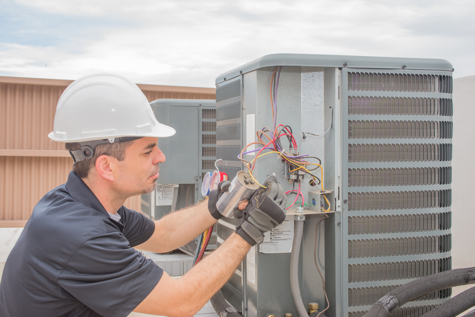 A man in blue overalls inspecting an air conditioner with a tool.